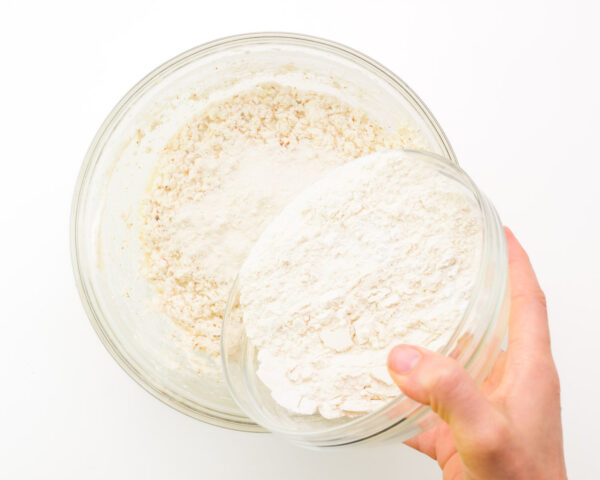 A hand holds a flour mixture, pouring it into a bowl with wet ingredients.