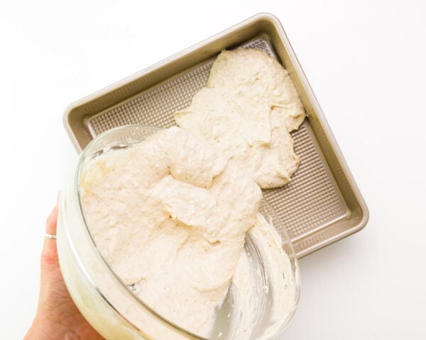 A hand holds a bowl, pouring cake batter into a baking pan.