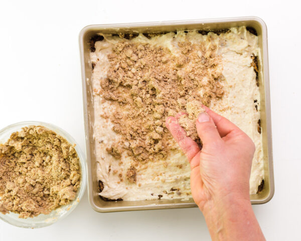 A hand is distributing crumble topping across cake batter in a baking pan.