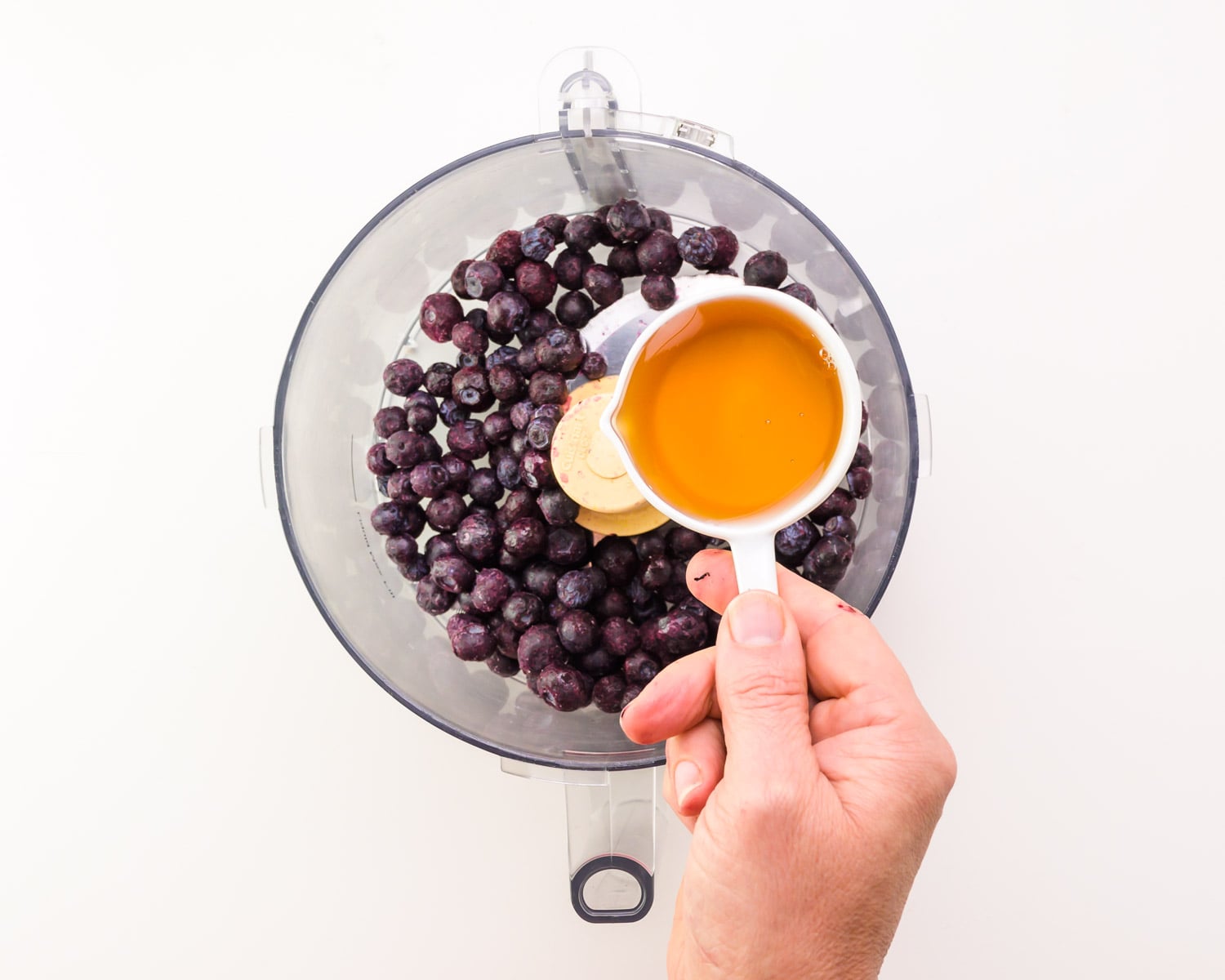Maple syrup is being poured from a measuring cup into a food processor with blueberries.