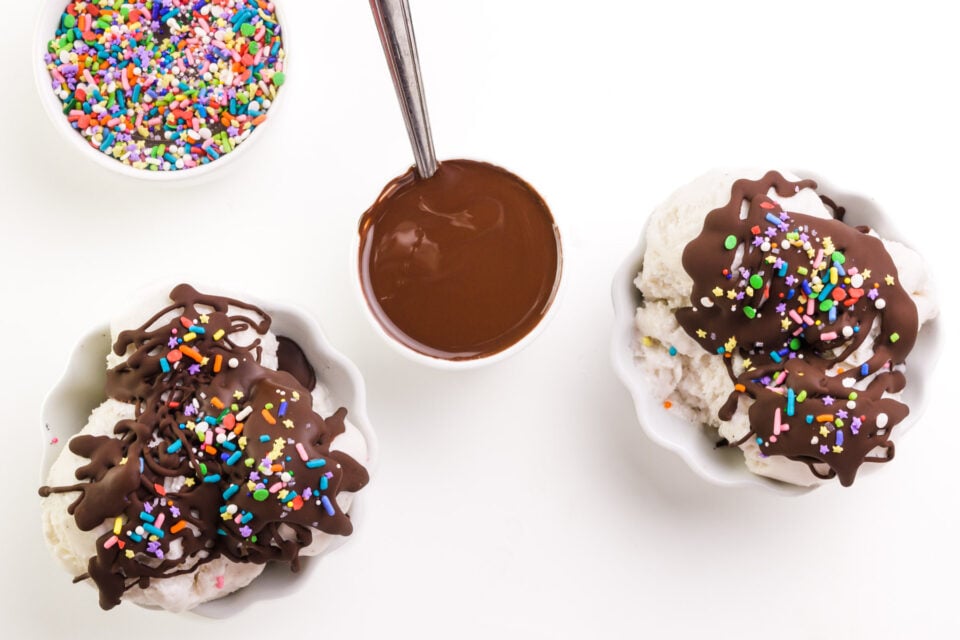 Looking down on a bowl of melted chocolate topping sitting next to bowls of ice cream with chocolate topping and sprinkles.