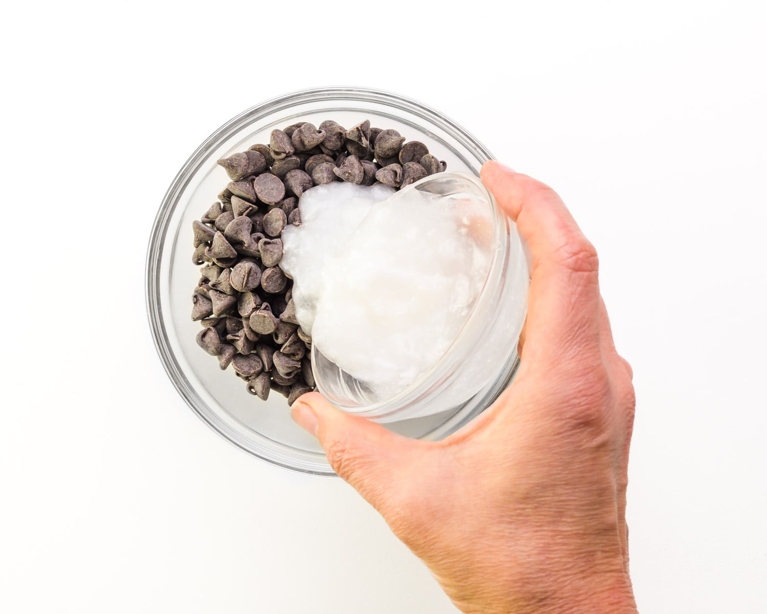 A hand holds a bowl pouring coconut oil into another bowl with chocolate chips.