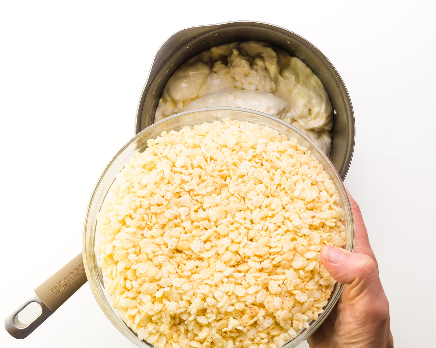 A large bowl of rice crispy cereal is being poured into a saucepan with melted marshmallow mixture.