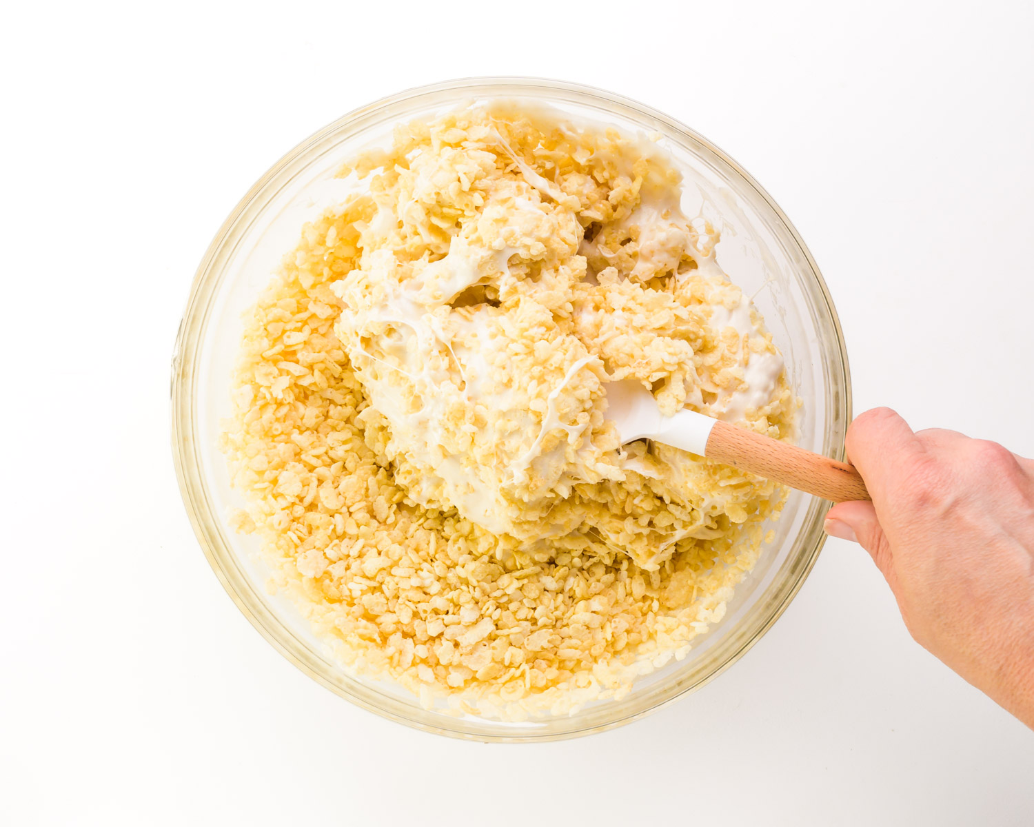 A hand holds a spatula stirring cereal into a marshmallow mixture.