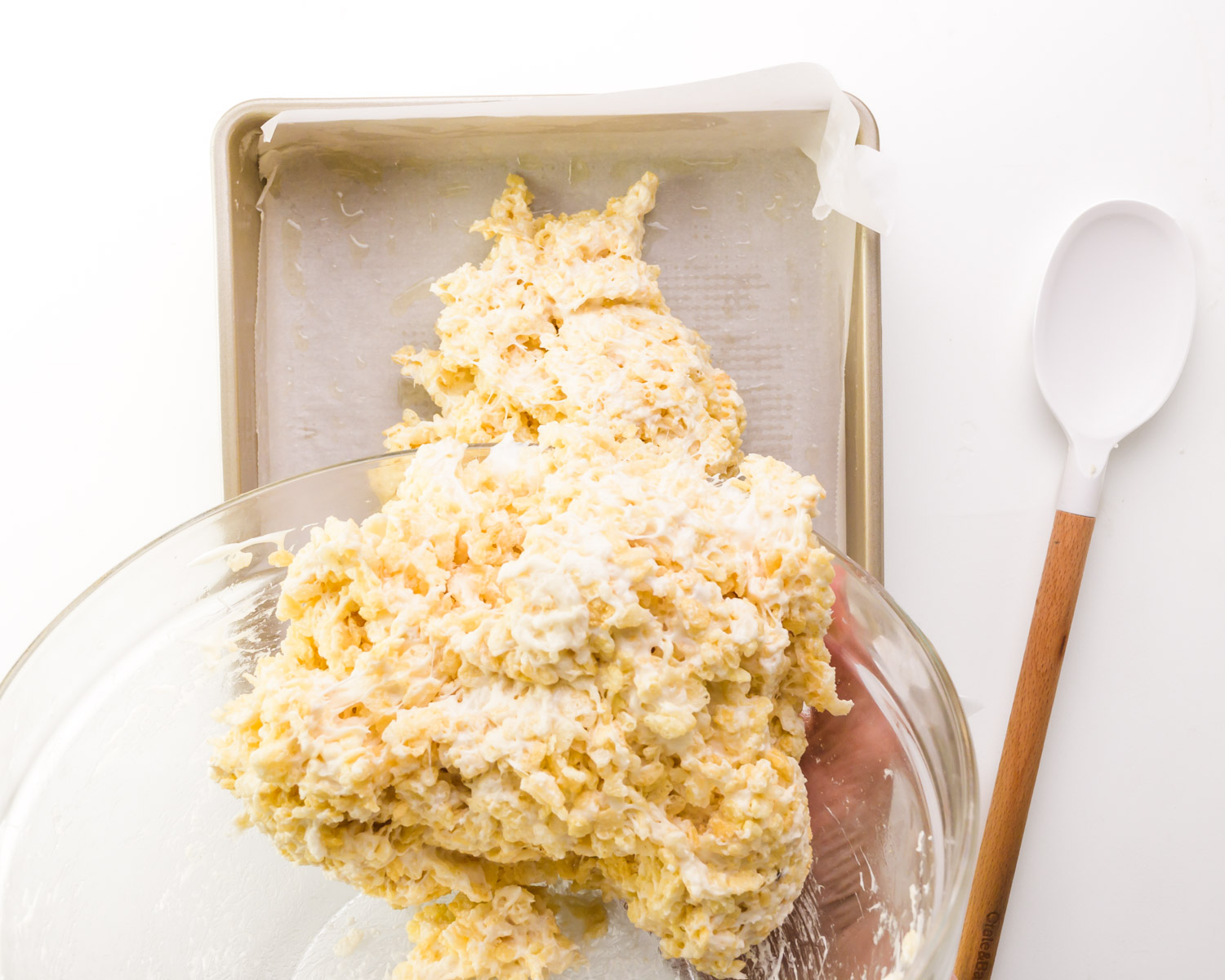 Marshmallow cereal mixture is being poured into a square baking pan. A spatula sits beside it.