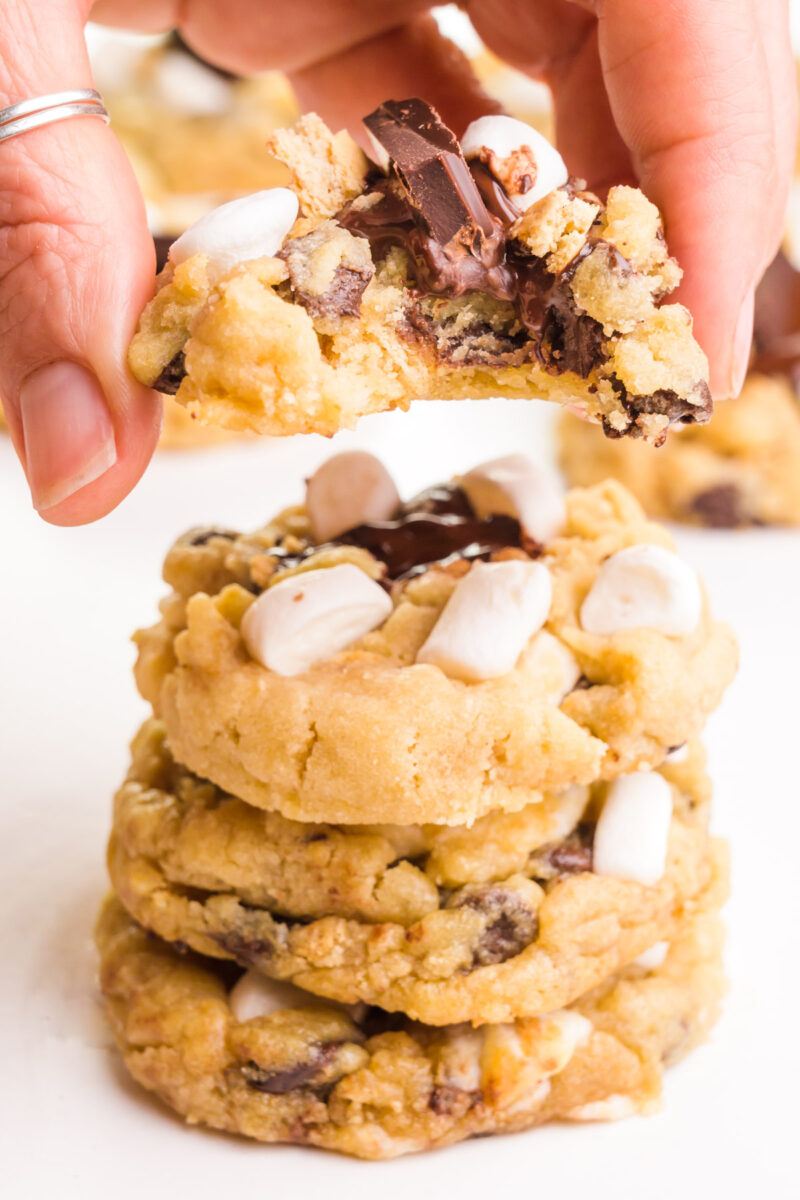 A hand holds a cookie with a bite taken out, hovering over a stack of the cookies.