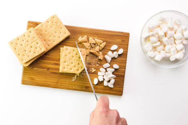 A hand holds a knife chopping graham crackers and marshmallows on a cutting board.