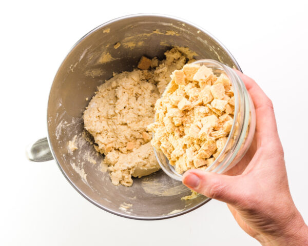 A hand holds a bowl of chopped graham crackers, pouring them into a bowl with cookie batter.