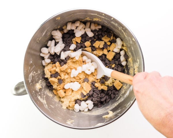 A hand stirs marshmallows and chocolate chips into cookie dough.