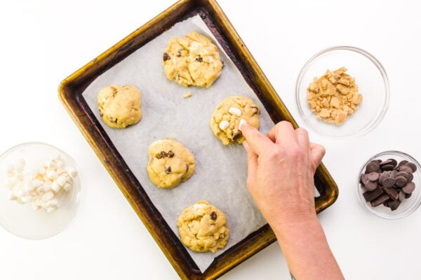 A hand presses marshmallows into cookies. There are bowls of chocolate chips, marshmallows, and chopped graham crackers next to the pan.