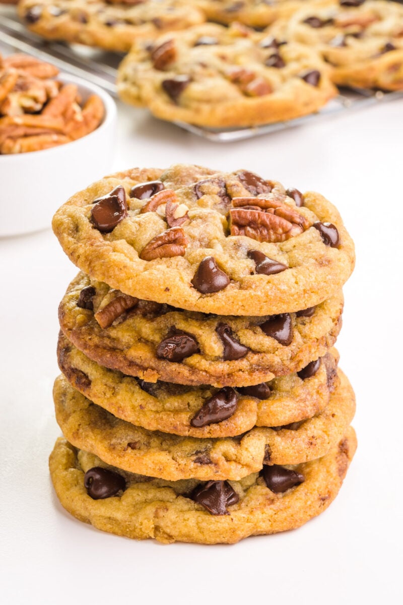 A stack of chocolate chip pecan cookies sits in front of a bowl of pecans and more cookies in the background.