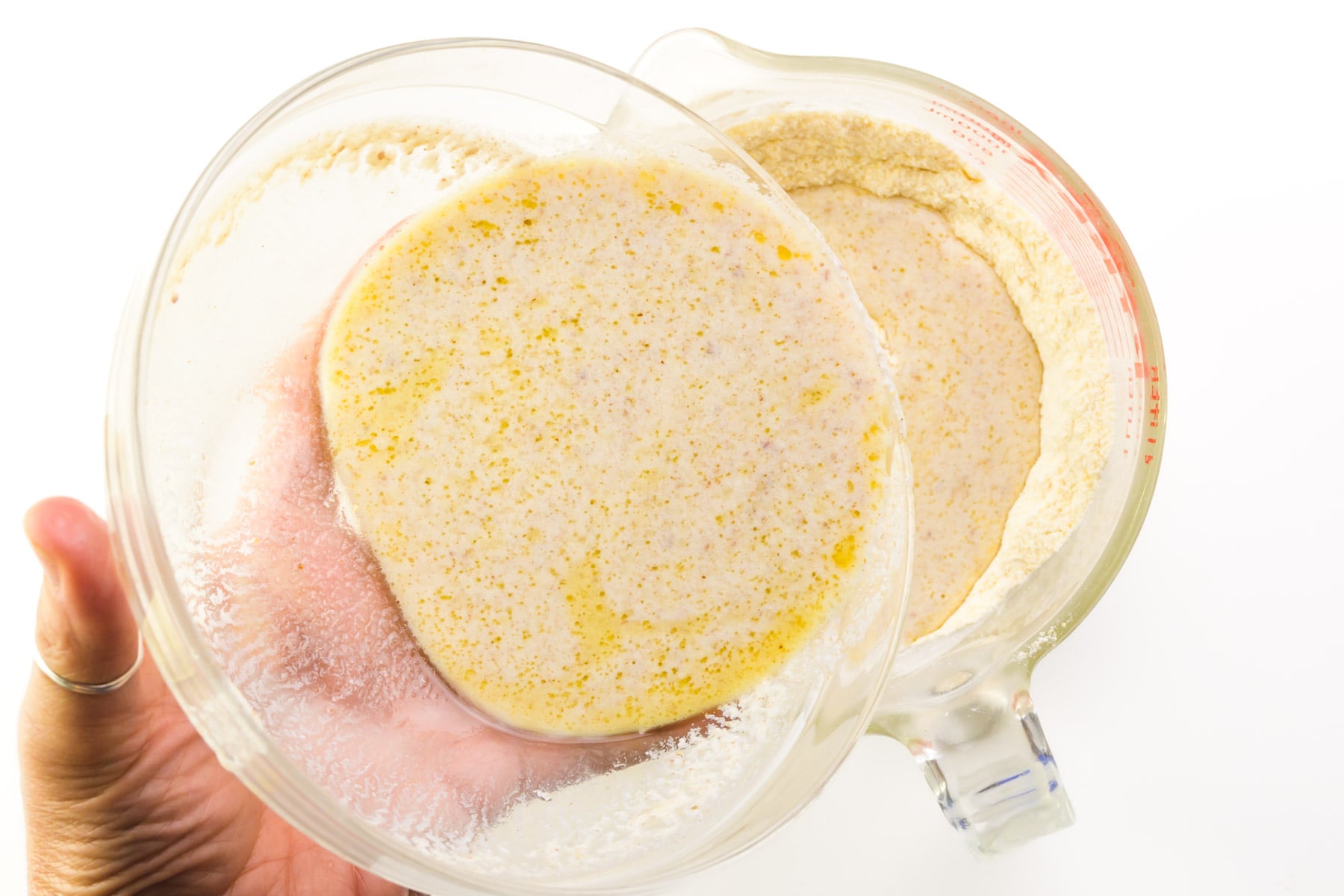 A hand holds a bowl of liquid ingredients, pouring it into a bowl with a cornstarch and flour mixture.