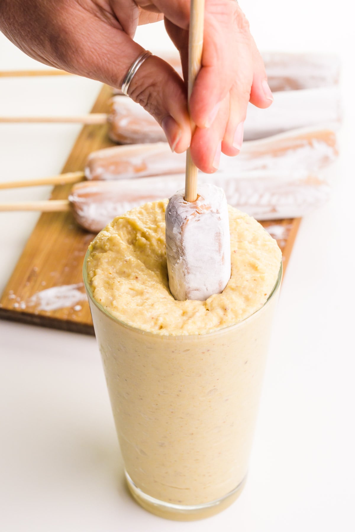 A hand holds a hot dog on a stick, dipping it in a tall glass full of cornbread batter. There is a cutting board in the background with more hot dogs to be dipped.