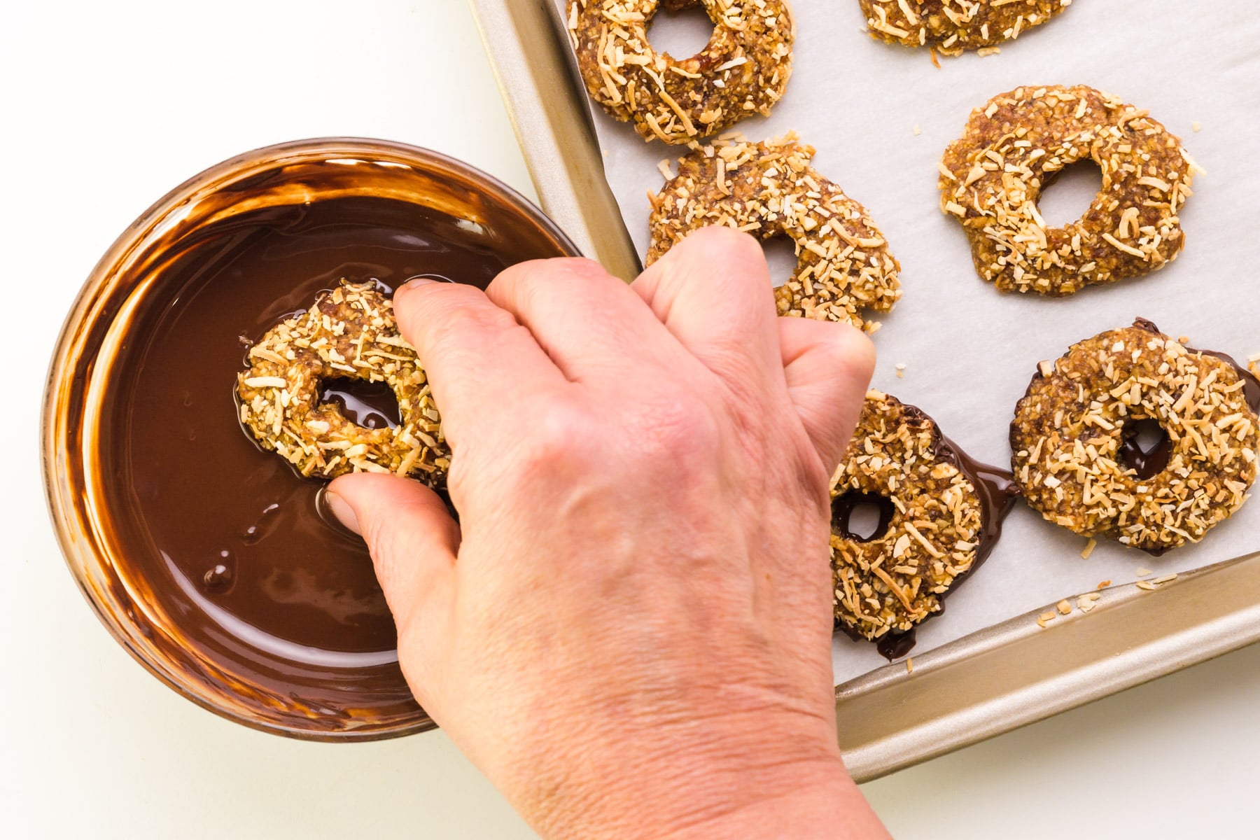 A hand dips a cookie in melted chocolate. A pan with more cookies sits beside it.