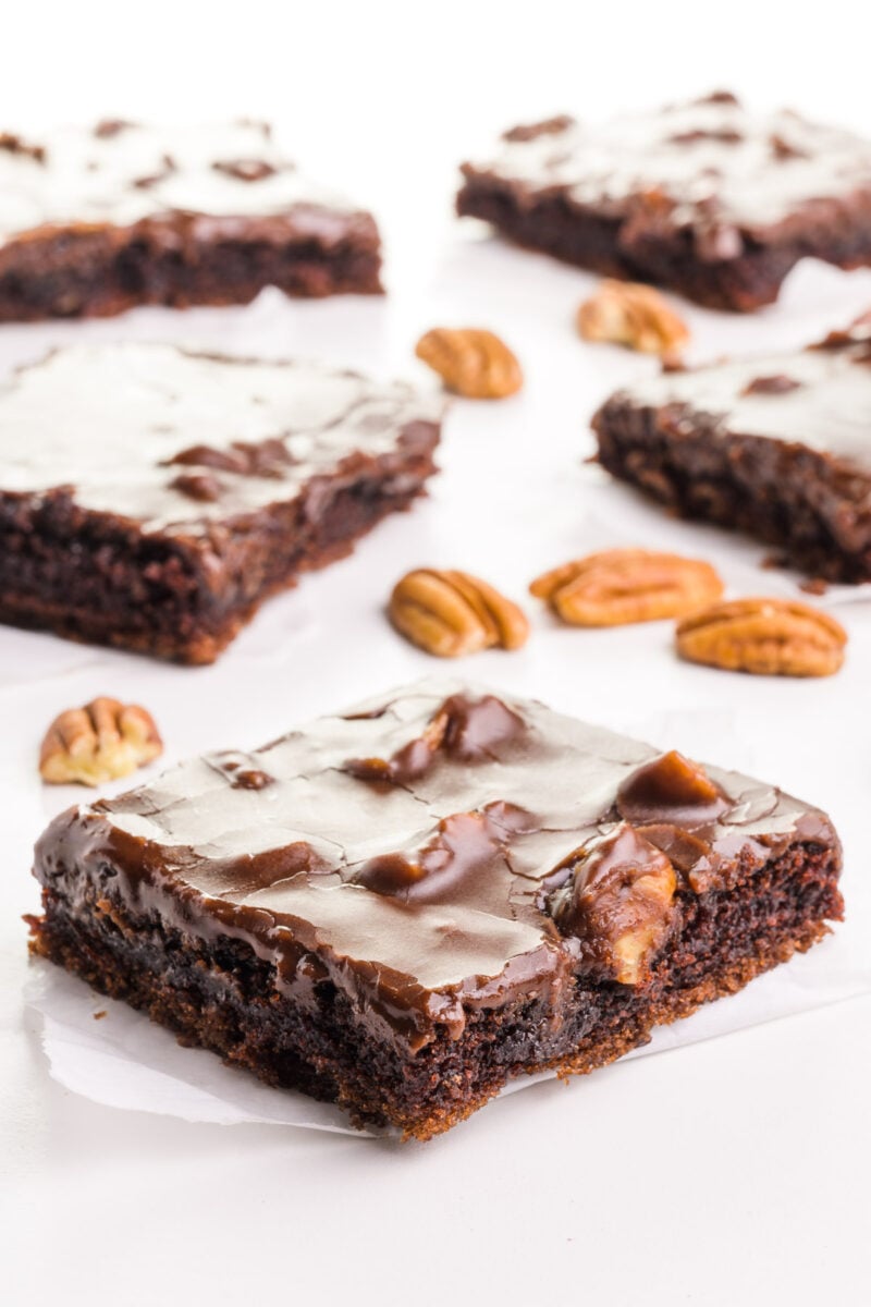 Several slices of vegan chocolate sheet cake sit on a white table, with pecans spread beside them.