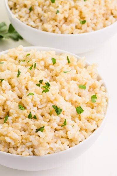 Two bowls of cooked brown rice sit next to sprigs of parsley.
