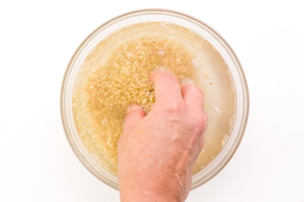 A hand reaches into a bowl with uncooked rice and water, stirring the rice.
