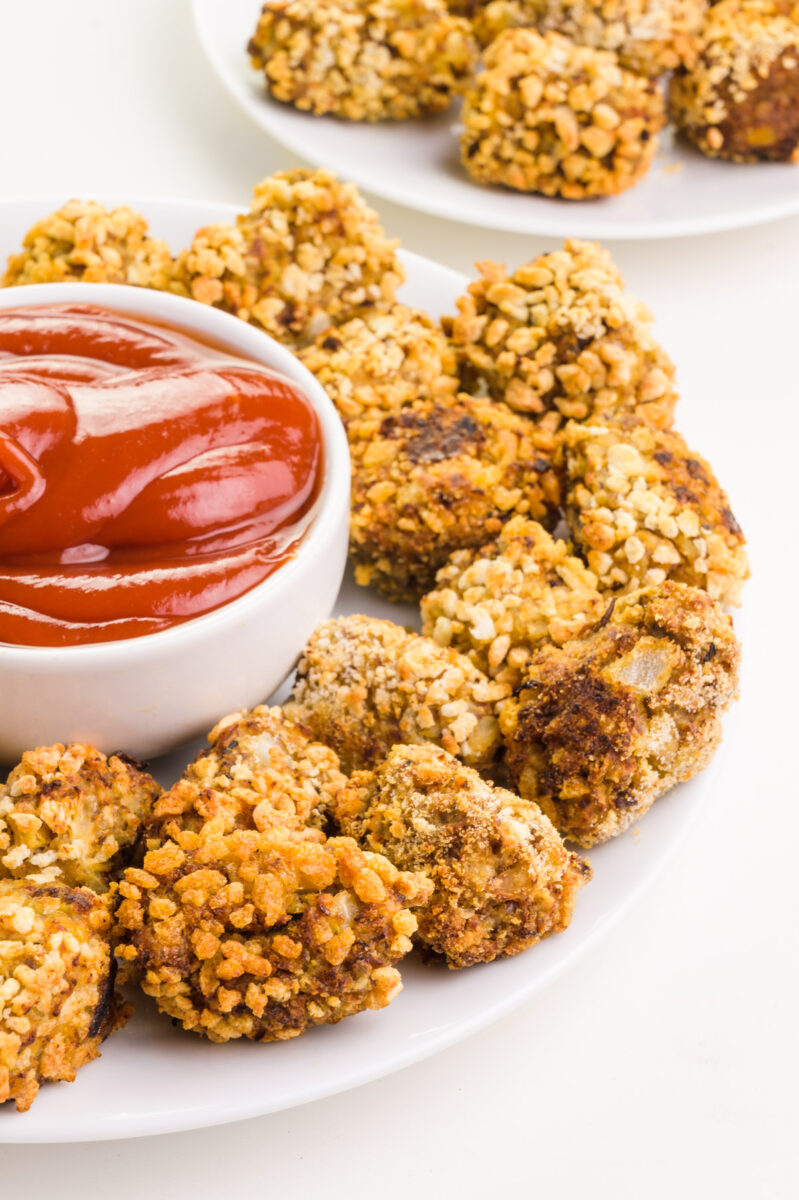 Cheesy cauliflower tots sit on a plate next to a bowl of ketchup. There are more tots in the background.