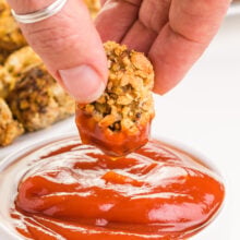 A hand holds a cauliflower tot after dipping it in a bowl of ketchup. There are more tots in the background.
