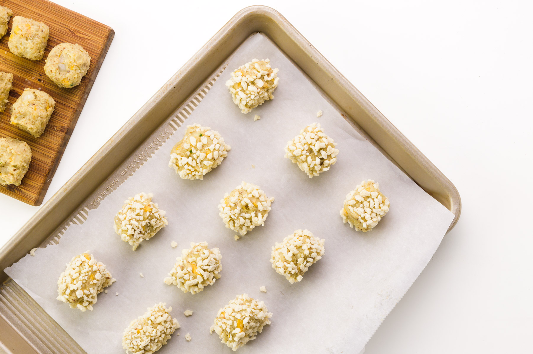 Tater tots are on a baking sheet lined with parchment paper. There are more tots on a cutting board beside the pan.