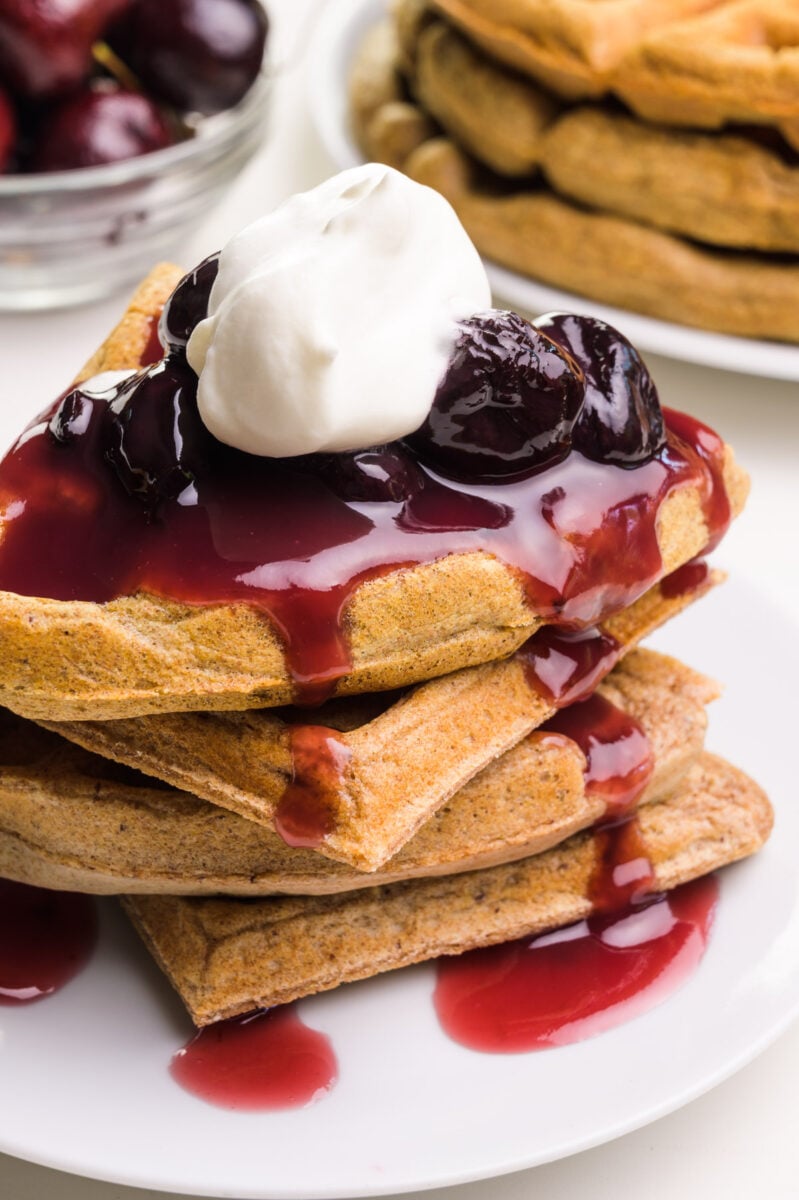 Waffles are smothered in cherry sauce and a dollop of whipped cream. There are more waffles and fresh cherries in the background.