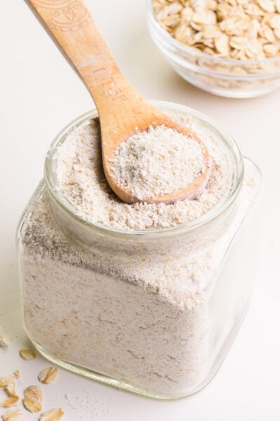 A wooden spoon sits in a bowl of oat flour. There is a bowl of oats in the background.