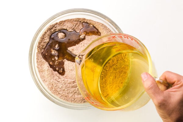 A hand holds a pyrex dish full of liquid, pouring it into a bowl with a cocoa powder mixture.