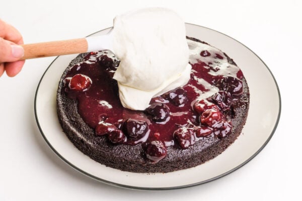 A spatula drops a dollop of whipped cream over cherry sauce on a chocolate cake.