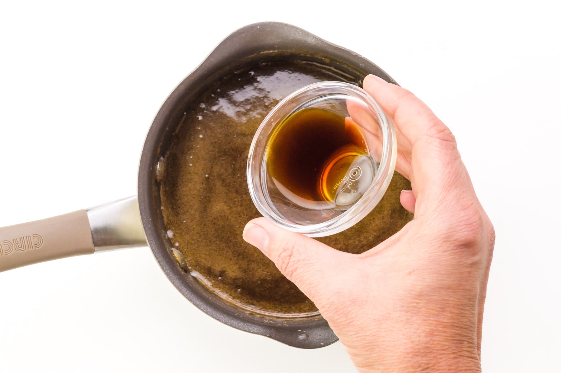 A hand holds a bowl of vanilla, pouring it into a saucepan with other ingredients.