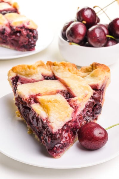 A slice of vegan cherry pie sits on a plate with a fresh cherry next to it. There is a bowl of cherries and another slice of pie in the background.