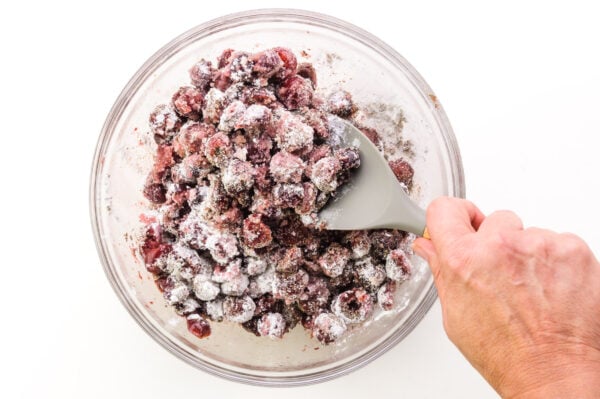 A hand holds a spatula, stirring cherry pie filling.