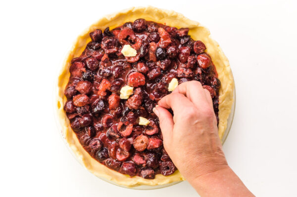 A hand places butter cubes over a fruit pie filling.