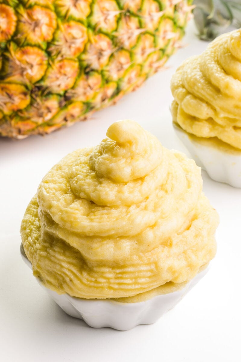 A bowl of vegan dole whip sits in front of a pineapple and another bowl of dole whip.