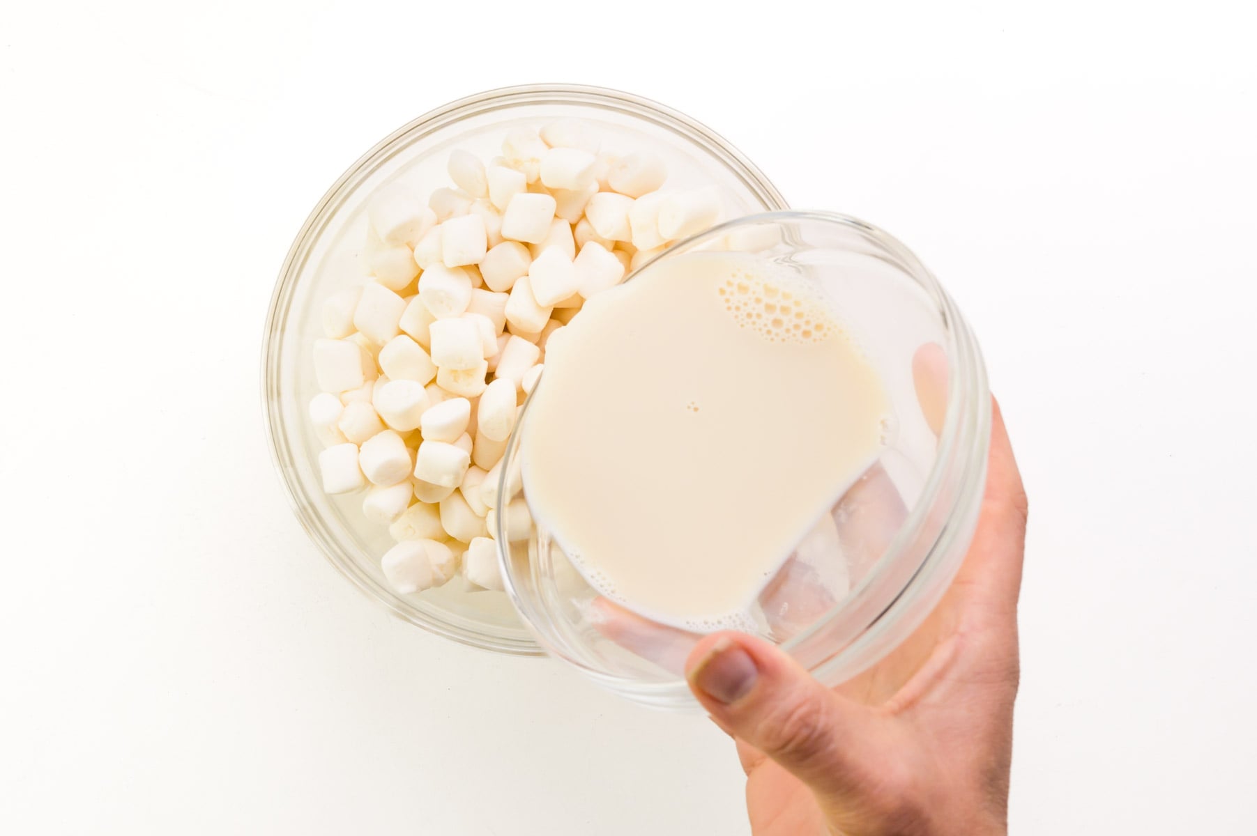 A hand holds a bowl of soy milk, pouring it into a bowl of marshmallows.