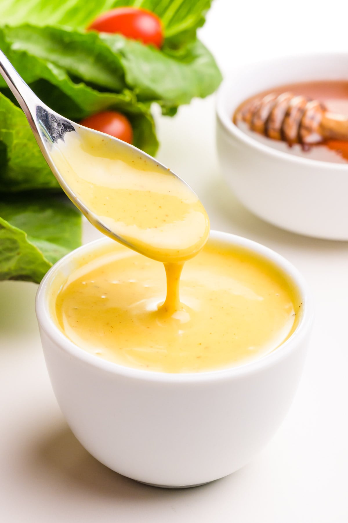 A spoon with vegan honey mustard dressing hovers over a bowl of it. There is a salad and a bowl with honey in the background.