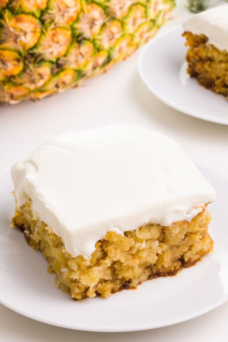 A slice of cake on a plate sits in front of a whole pineapple on its side and another slice of cake.