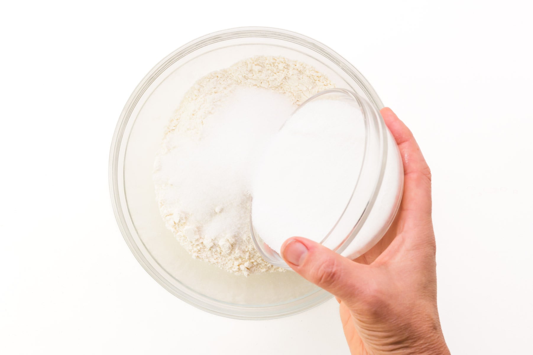 A hand pours a bowl of granulated sugar into a bowl of flour.
