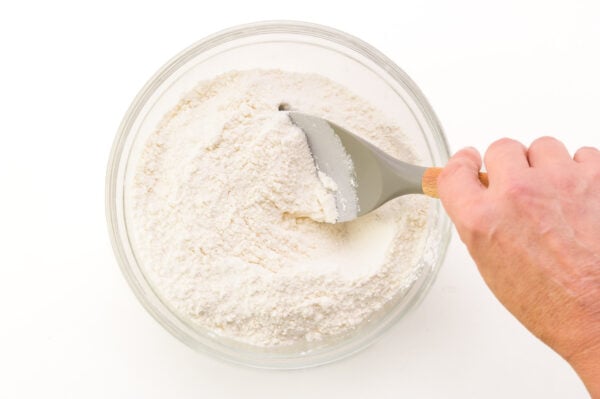 A hand holds a spatula, stirring together flour ingredients in a bowl.