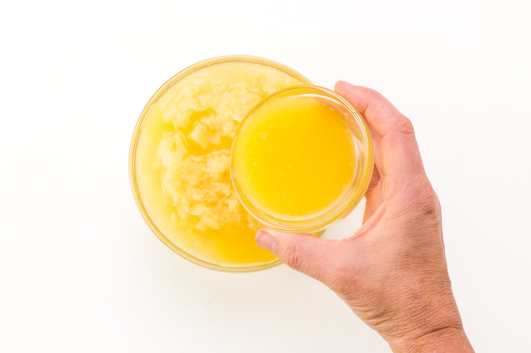 A hand holds a bowl of melted butter, pouring it into a bowl with crushed pineapple.