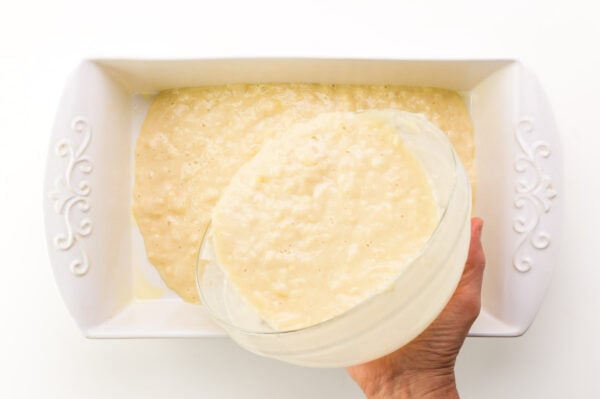 Cake batter is being poured into a white baking dish.