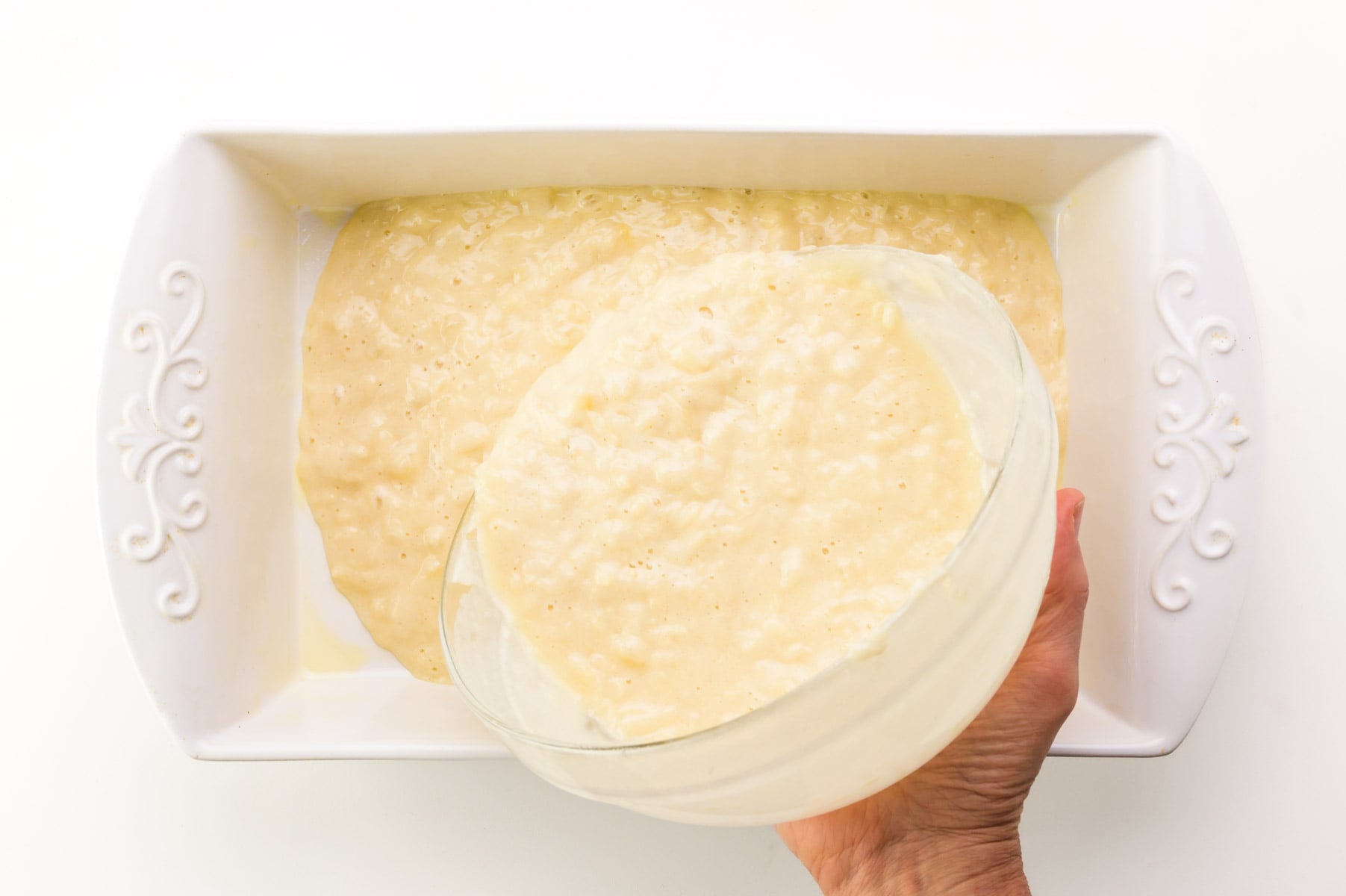 Cake batter is being poured into a white baking dish.