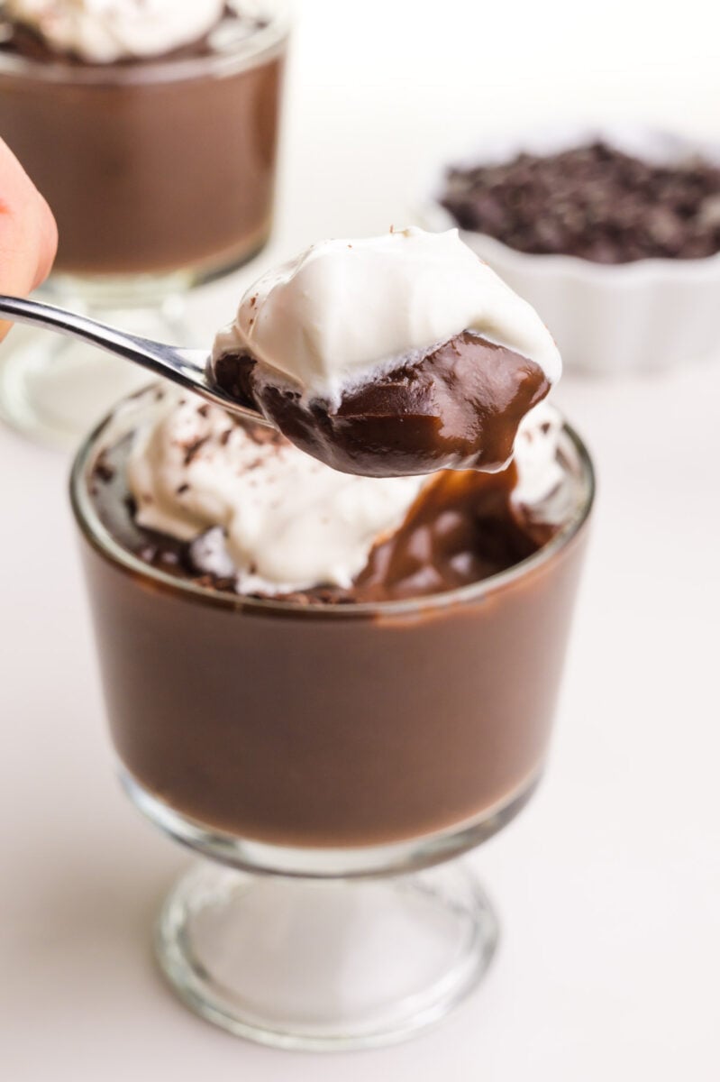 A hand holds a spoonful of vegan chocolate pudding topped with whipped cream. It hovers over a serving dish full of pudding.