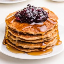A stack of oatmeal pancakes sit on a plate with maple syrup drizzling over the sides. There is a dollop of blueberry sauce on top.