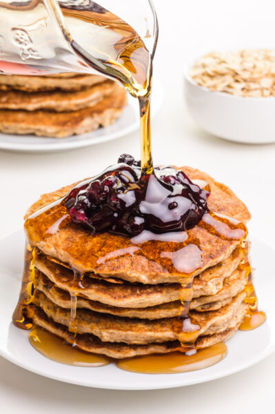 Vegan oatmeal pancakes are on a plate with blueberry sauce on top. Maple syrup is being poured on top. There is another plate of pancakes and a bowl of oats in the background.