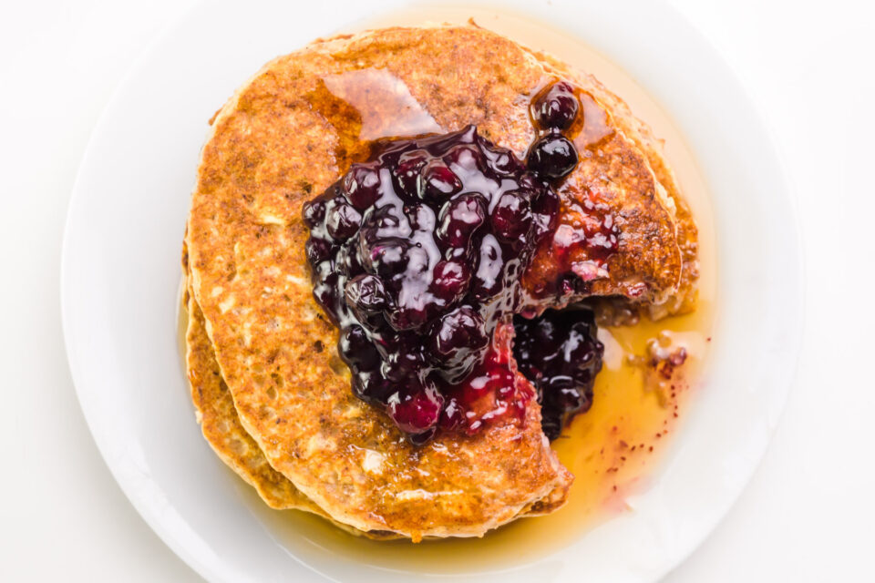 Looking down on pancakes on a plate with a triangle bite taken out, revealing syrup and blueberries on the plate.