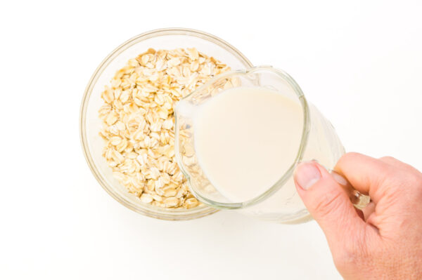 A hand holds a pitcher of milk, pouring it into a bowl of oats.