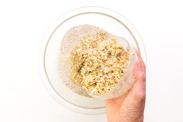 A hand holds a bowl of moistened oats, pouring them into a bowl with flour.