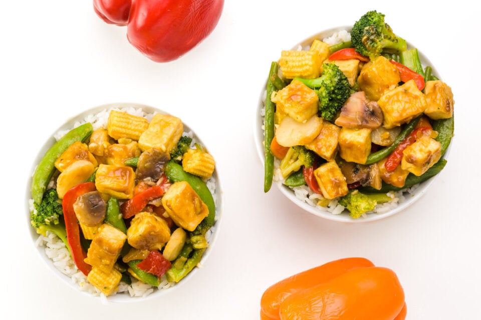 Looking down on two bowls of stir-fried tofu. There is a red and yellow bell pepper next to the bowls.