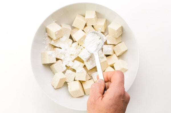 A hand holds a measuring spoon of cornstarch, sprinkling it over tofu cubes in a bowl.