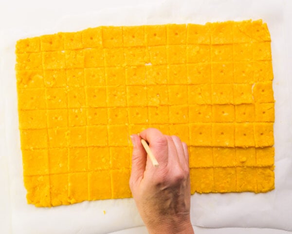 A hand holds a chopstick, pressing dots into squares of orange dough.